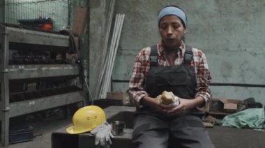 Medium long of female Hispanic plant worker eating sandwich, drinking coffee, sitting at factory in afternoon, having lunch, smiling