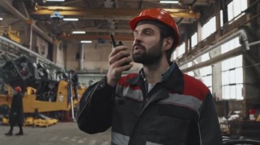 Medium close-up of Caucasian bearded male plant manager talking into walkie talkie at factory in afternoon