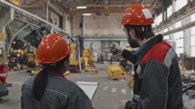 Sakallı beyaz adam ve siyah şapkalı ve üniformalı genç Afro-Amerikan kadın özel araç fabrikasında dikiliyor, konuşuyorlar, pano ve telsiz taşıyorlar.
