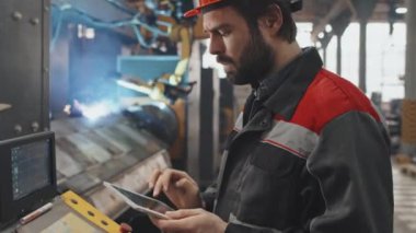 Tilting down slow motion of male Caucasian engineer in orange helmet and uniform using tablet computer for configuration of special equipment at factory