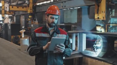 Turuncu kasklı, üniformalı, sakallı beyaz adamın tablet bilgisayar kullanarak belden yukarı portresi. Sonra da fabrikada kaynak yaparken kameraya bakıyor.