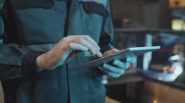 Tilting up of bearded Caucasian man in orange helmet and uniform using tablet computer, standing in foreground of welding at factory