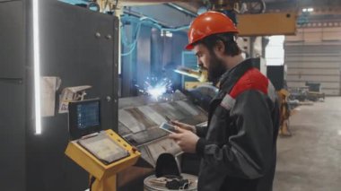 Turuncu başlıklı ve üniformalı beyaz erkek mühendisin fabrikadaki özel ekipmanların yapılandırılması için tablet bilgisayar kullanışının orta boy yan görüntüsü.