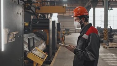 Beyaz tenli turuncu kasklı, maskeli ve üniformalı erkek teknisyenin tablet bilgisayar kullanarak, tesisteki özel ekipmanları kontrol ederken,