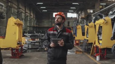 Kafkas sakallı, turuncu şapkalı işçinin orta boy takibi. Ağır makine üretim tesisinde yürüyor, tablet bilgisayar kullanıyor.