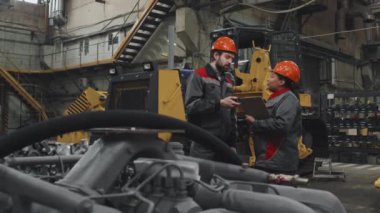 Orta uzunlukta beyaz erkek mühendis ve kadın Afro-Amerikan yönetici. Ağır makine üretim fabrikasında dikilip konuşuyorlar.
