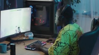 Medium side shot of African American male teenager wearing yellow t-shirt and headset sitting in his room at home, playing shooter video game on computer, failing and reacting with disappointment