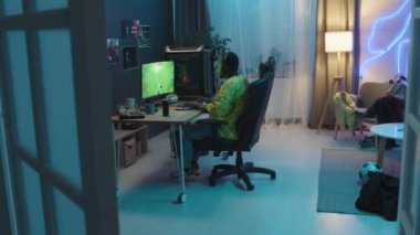 Wide shot of young African American man sitting in neon-lit apartment in front of professional computer screen, playing shooter videogame, winning level and pumping his fists in joy