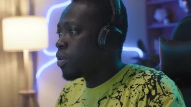 Medium close-up shot of teenage black male gamer with headphones sitting in computer chair, anxiously looking at screen, throwing up hands and booing in dismay, blue neon light in background
