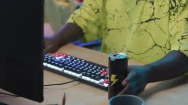 Medium close-up tilting shot of hands and face of young black man in headphones sitting in front of computer, looking at screen, pressing buttons on keyboard, and consuming canned energy drink