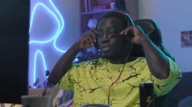 Medium shot of teenage black male gamer sitting in front of computer in neon-lit room at home, getting ready to play online videogame, putting on headset and talking into microphone