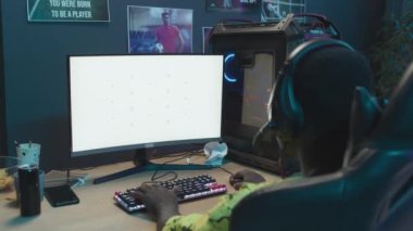 Over-the-shoulder shot of African American teenager with headset sitting at home in front of gaming computer with empty white screen and playing. Suitable as mockup, chromakey template