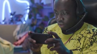 Medium close-up shot of face of happy young African American gamer sitting in room and playing videogame on smartphone, scoring, cheering and clenching fist with joy. Bright neon light in background