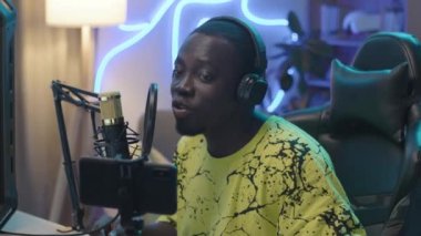 Medium close-up shot of African American guy with headphones sitting in front of computer, talking into microphone, looking at smartphone while answering questions from subscribers during livestream