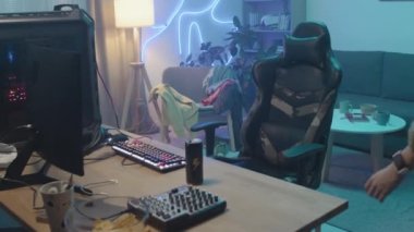 Lockdown medium shot of young Asian woman with short hair and glasses walking up to desk, sitting down in front of gaming computer, putting on headset and starting to play and speak into microphone