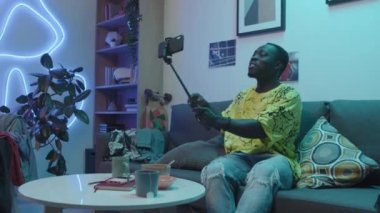 Full shot of young African American student sitting on couch, holding smartphone on selfie stick and turning around while talking to friend on video call and showing him his new room