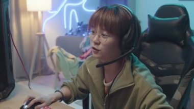 Medium close-up shot of young Chinese female gamer with short hair, glasses and headset sitting in front of computer, chatting into microphone, smiling and playing video game
