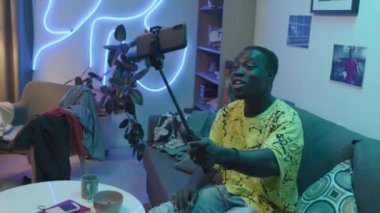 High-angle medium shot of young black guy sitting on couch in his neon-lit room at home, holding smartphone on selfie stick, looking at camera, talking expressively and gesticulating