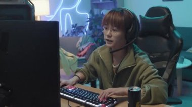 Medium shot of Asian teenage girl sitting in front of computer at home, playing intensive shooter game on keyboard, winning, clapping, cheering and raising hands in joy and triumph