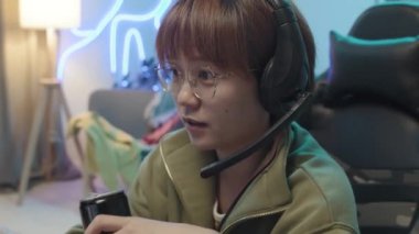 Medium close-up shot of face of young Asian female gamer sitting in her room with blue neon lighting, sipping energy drink from can, and talking into microphone with playing companions