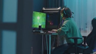 Full side shot of unrecognisable gamer sitting at desk late at night and playing FPS online videogame on computer, firing bullets from gun and hitting targets