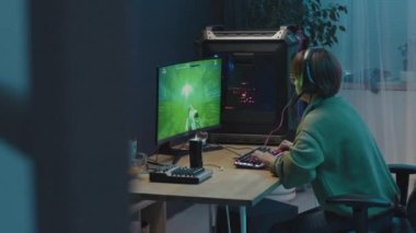 Medium shot of young Chinese woman sitting in front of computer at home, playing first-person shooter video game, completing level, taking off headphones, cheering, standing up and walking away