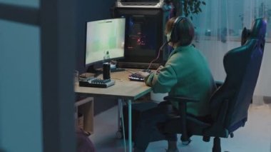 Full side shot of female gamer sitting in room at night, playing first-person shooter with intensive gameplay on computer, completing level, taking off headset and cheering with delight at achievement