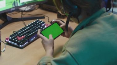 Over--the-shoulder shot of anonymous gamer sitting at desk in front of computer with videogame on, and playing mobile game on smartphone, with green screen. Suitable as chromakey template, copy space