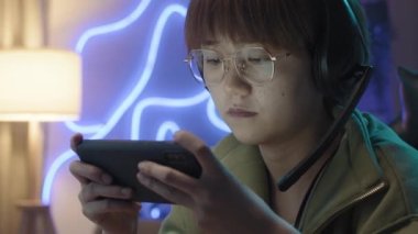 Close-up shot of nerdy young Chinese woman with headset, short hair, glasses and braces on teeth playing mobile game on smartphone, winning and cheering with delight, blue neon light in background