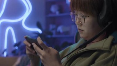 Close-up side shot of face and hands of young Chinese woman in glasses, with headset, reclining in chair and enthusiastically playing mobile game on smartphone, with dim blue neon light in background