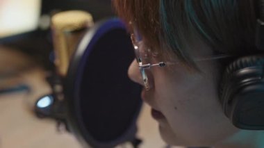 Close-up side shot of face of male or female Asian person in glasses talking into microphone while playing videogame on computer and livestreaming to subscribers