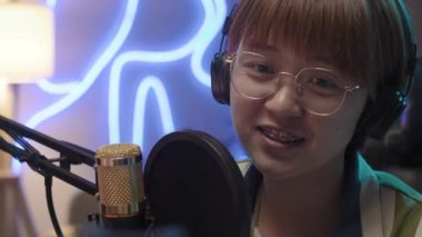 Close-up shot of smiling young Asian woman in glasses and headphones talking into studio microphone and looking confidently at camera