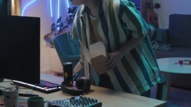 Medium lockdown shot of young Chinese female gamer walking up to desk, sitting down on chair, putting feet up, and eating takeaway meal with chopsticks while watching action on screen