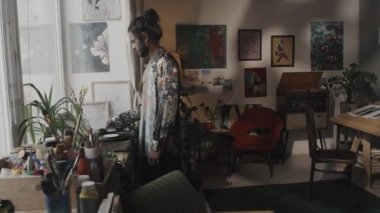 Young male Indian artist with long dark hair in bun and unbuttoned floral printed silk shirt turning on music on retro vinyl record player and starting work in creative messy studio