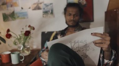 Stylish young male Indian fashion designer looking at sketch and thinking, sitting in red armchair at creative workshop with colorful paintings on walls