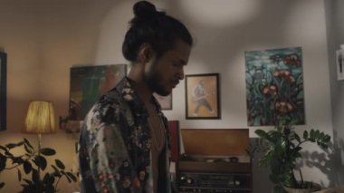 Tilt down shot of talented young male Indian fashion designer in unbuttoned floral printed silk shirt looking at clothes sketches and inspiration ideas in creative workshop