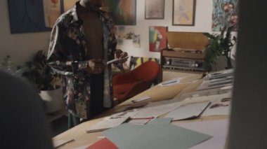 Male Indian fashion designer standing by wooden table with clothes sketches and cutting out paper pattern sample with scissors, working in creative messy studio