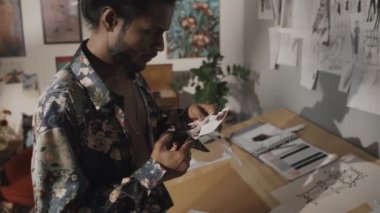 Talented Indian fashion designer cutting out small print sample with scissors, working in creative mess in retro style studio