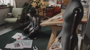 Young male Indian fashion designer sitting on floor in creative messy studio, cutting inspirational pictures for mood board