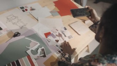 From above of handsome fashion designer with painted black nails taking photos of inspirational mood board and clothes sketches on wooden worktable in studio
