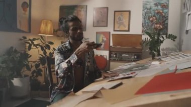 Young Indian male fashion designer in floral printed silk shirt with long dark hair in bun taking photos pf clothes sketches at worktable in messy colorful studio