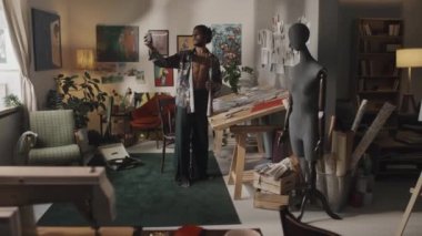 Genç Hintli erkek moda tasarımcısının düğmeleri iliklenmemiş ipek gömlekli, akıllı telefonuyla video blogu kaydeden yaratıcı retro tarzı dağınık çalışma alanı.