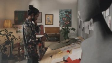 Medium shot of young handsome Indian fashion designer listening to music in earphones while creating new collection in creative retro style messy workshop