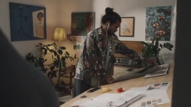 Young handsome Indian fashion designer in earphones cutting out pattern from grey fabric on wooden worktable in creative workshop