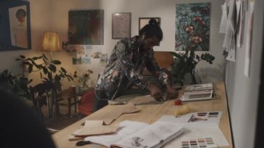 Medium shot of young male Indian fashion designer cutting fabric with paper pattern at wooden worktable in atelier