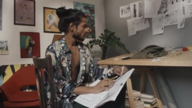 Eccentric young Indian fashion designer with long dark hair in bun wearing unbuttoned floral printed silk shirt drawing in sketchbook while working on new collection in messy atelier