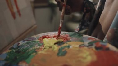 Unrecognizable male artist with bare torso mixing colors on artist palette while painting on canvas in studio