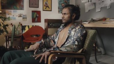 Portrait of handsome young Indian designer or artist in unbuttoned floral printed silk shirt with painted black nails sitting in armchair in creative messy workshop looking at camera