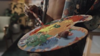 Unrecognizable tanned male artist with bare torso using artist palette while painting on canvas in studio