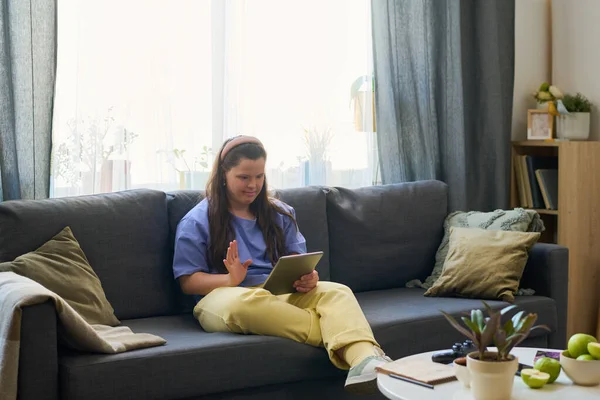 Girl with Down syndrome waving hand during communication in video chat while sitting on sofa and greeting person on tablet screen